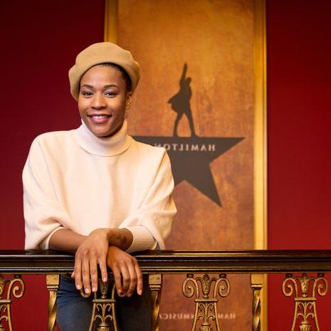 A woman wearing a tan beret and a beige sweater leans on a railing inside the Hamilton theater.