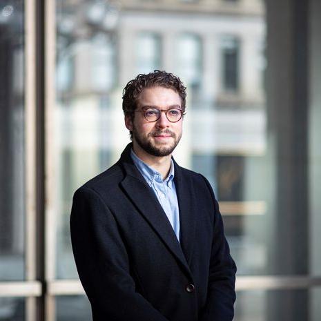 A man with a beard and wearing a suit poses in front of a window.